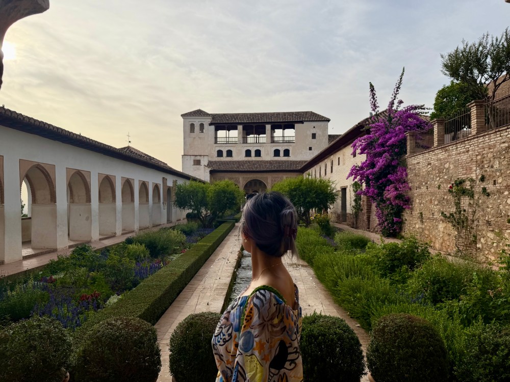 Person gazing at a historic building with gardens and purple flowers.
