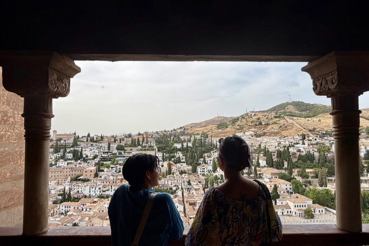Two people view a scenic cityscape with hills from a balcony.