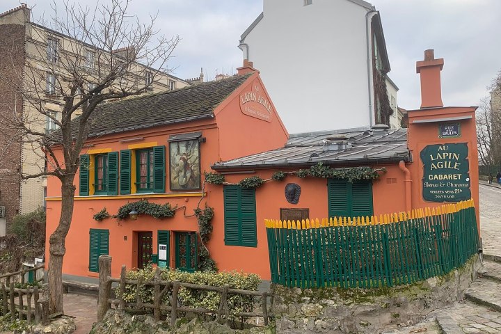 Historic red building with green shutters and a sign for 'Le Lapin Agile' in a cobblestone setting.