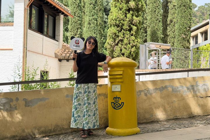 Person in sunglasses holds a sign next to a yellow post box, with trees and buildings in the background.