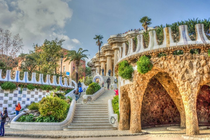 Colorful park with mosaic stairs, arches, and lush greenery, people walking and sitting.