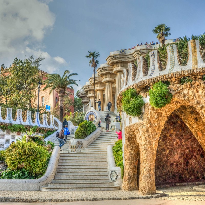 Colorful park with mosaic stairs, arches, and lush greenery, people walking and sitting.