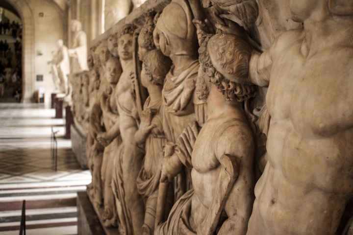 Side view of an ancient carved stone Roman sarcophagus depicting a detailed high-relief row of classical mythic figures and deities at the Louvre.