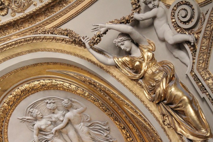 Low-angle view of an ornate, gilded neoclassical ceiling in the Louvre Museum featuring sculpted marble figures in relief and gold decorative leafing.